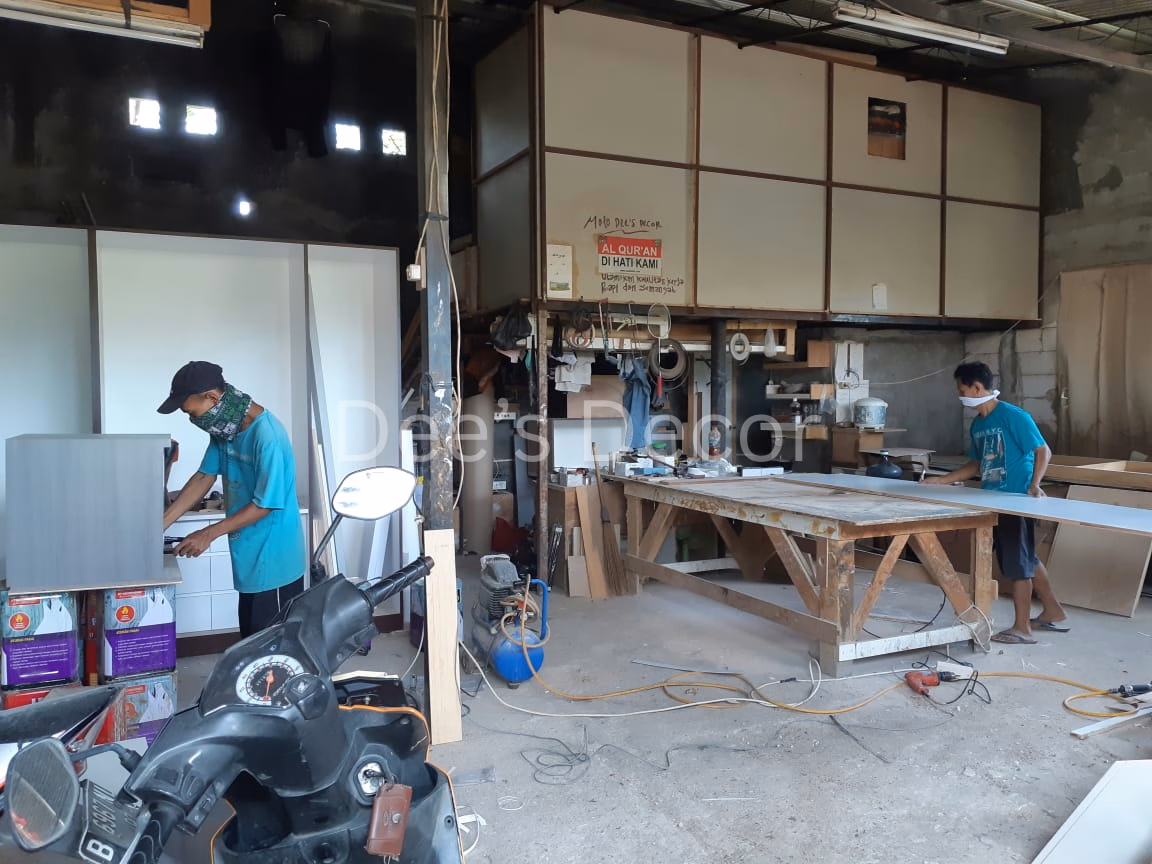 Workshop pembuatan furniture dengan dua tukang kayu sedang mengerjakan lembaran kayu di meja kerja besar di dalam bengkel sederhana.