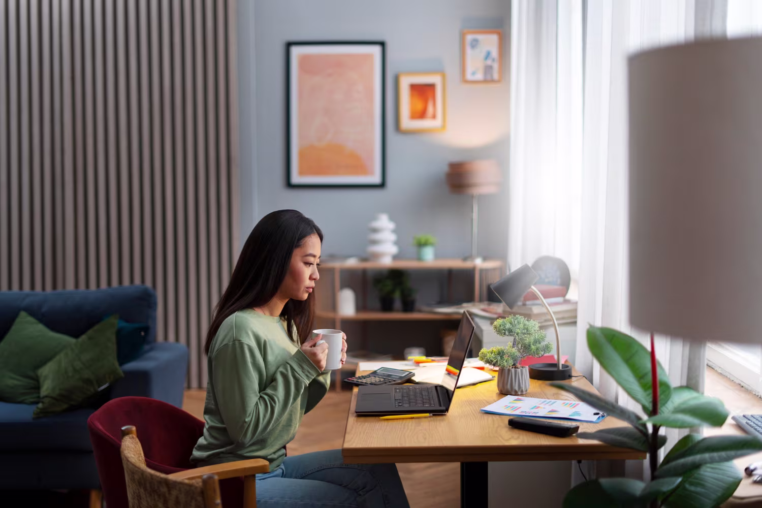 Wanita Asia bekerja dari rumah di meja kayu dengan laptop sambil memegang cangkir kopi di ruangan bergaya modern hangat