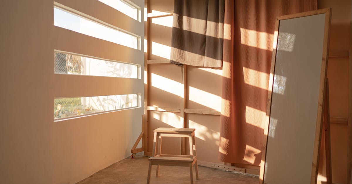 A minimalist room in Indonesia with warm sunlight, featuring a wooden stool, mirror, and curtains.