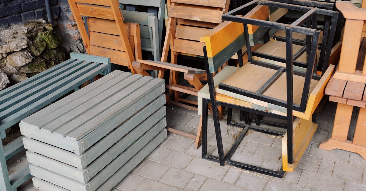 A mix of wooden benches, chairs, and metal frames stacked outside on a paved area.
