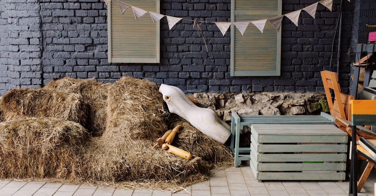 Body of mannequin without hands and legs placed on haystacks near wooden box and metal bench near black brick wall of mo