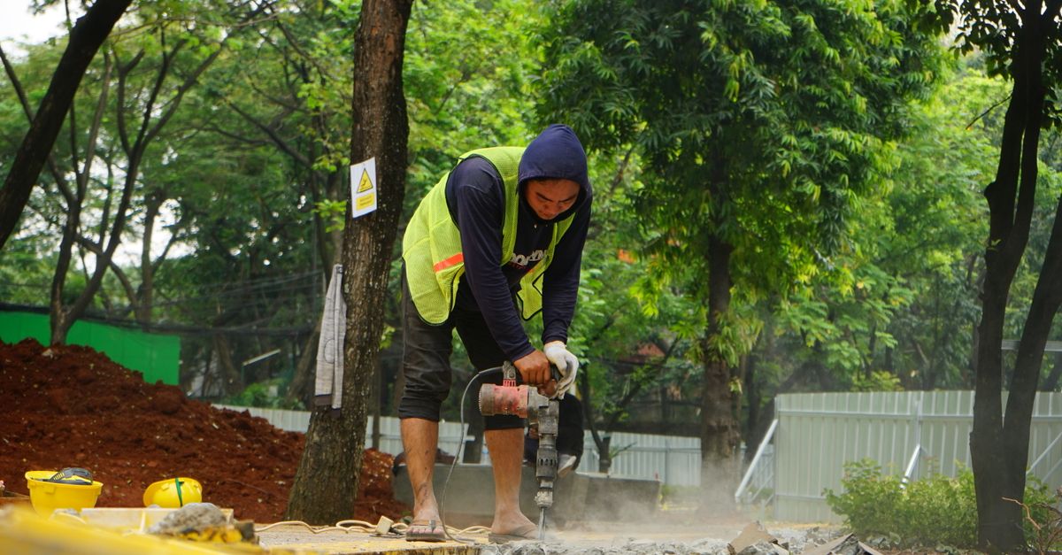 A construction worker in casual attire operates a drill on a site surrounded by greenery.