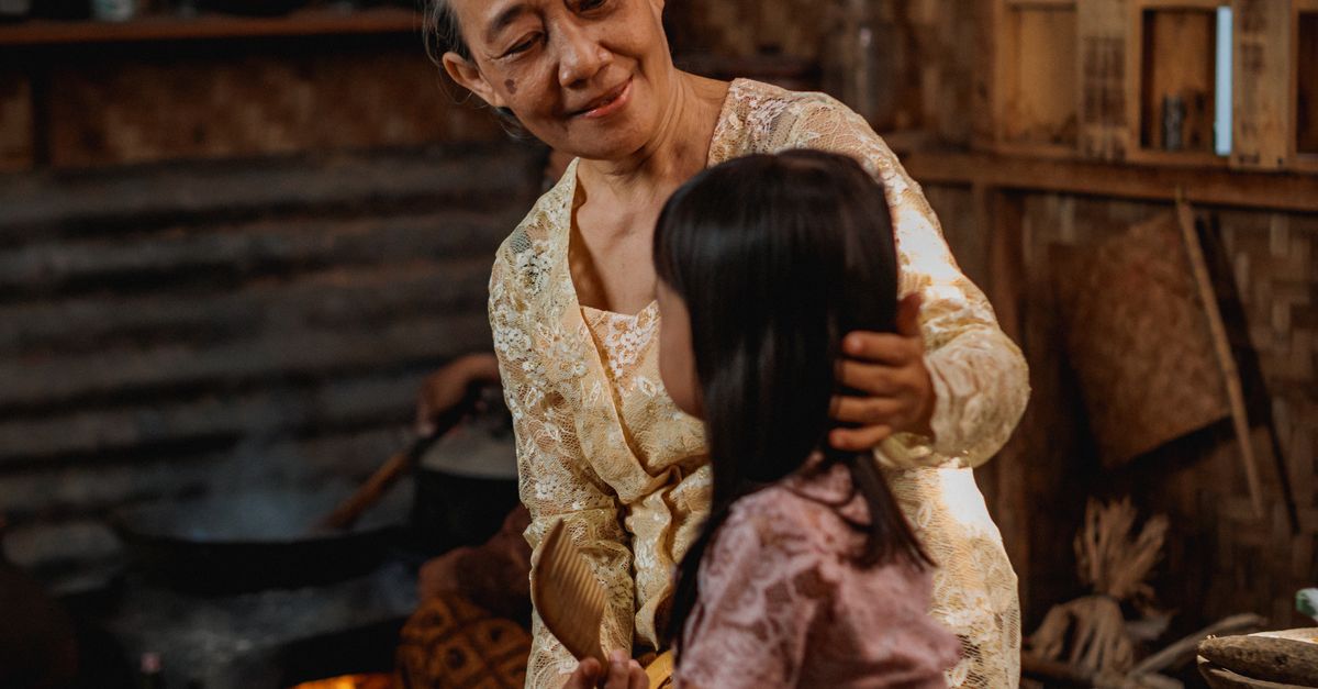 An elderly woman and a young girl share a touching moment in a traditional Indonesian bamboo kitchen.